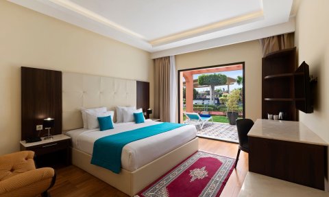 Ein modernes Hotelzimmer mit einem großen Bett, bunten Kissen und Blick auf einen gepflegten Gartenbereich.