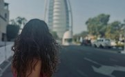 Eine junge Frau mit lockigem Haar trägt ein pinkes Oberteil und schaut auf ein modernes Gebäude in Dubai.