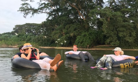 Fünf Personen entspannen auf Reifen im Wasser, umgeben von üppigem Grün und fliegenden Vögeln im Hintergrund.