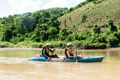 Zwei Personen im Kajak paddeln fröhlich auf dem ruhigen Nam Khan Fluss, umgeben von üppiger grüner Landschaft.