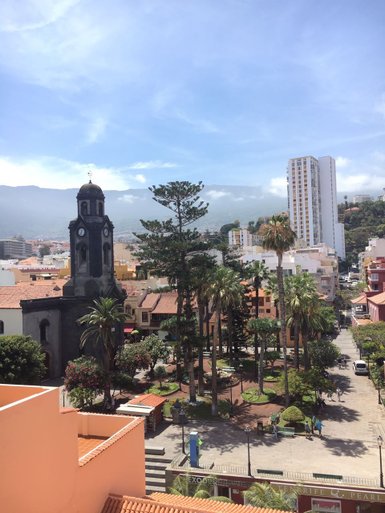 Ein Blick auf den lebhaften Platz in Puerto de la Cruz, umgeben von Palmen und bunten Gebäuden unter einem blauen Himmel.