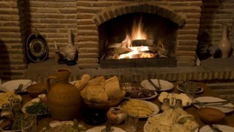 Ein rustikaler Tisch ist mit verschiedenen georgischen Gerichten und frischem Brot gedeckt, während ein Kamin im Hintergrund brennt.
