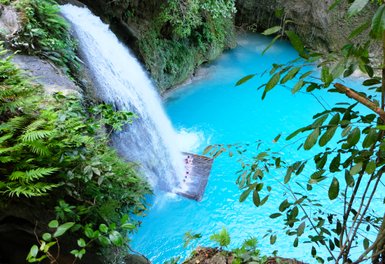 Ein Wasserfall stürzt in ein leuchtend blaues Wasserbecken, umgeben von üppigem Grün und tropischen Pflanzen.