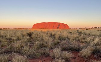 Ayers Rock strahlt in der Abenddämmerung, umgeben von sanften, grasbewachsenen Hügeln und dem klaren Himmel im Hintergrund.
