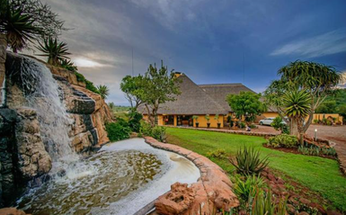Eine malerische Lodge mit einem Wasserfall und üppigem Grün, umgeben von tropischen Pflanzen und einem bewölkten Himmel.