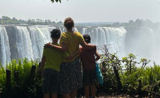 Eine Mutter umarmt ihre beiden Kinder, während sie die majestätischen Victoriafälle im Hintergrund bewundern.