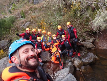Eine Gruppe von zehn Personen in Neoprenanzügen und Helmen posiert fröhlich am Ufer eines Gewässers.