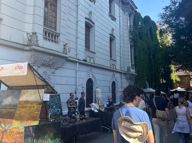 Ein belebter Markt mit Kunstständen vor einem eleganten, historischen Gebäude, umgeben von üppigem Grün.