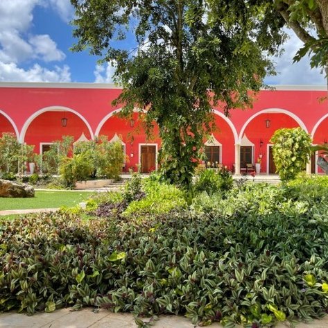 Ein lebendiger Garten mit üppigem Grün umgibt ein rotes Gebäude mit eleganten Bögen und Türen, unter einem strahlend blauen Himmel.
