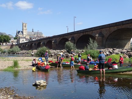 Eine Gruppe von Menschen in Kanus bereitet sich auf eine Kanutour an einem sonnigen Flussufer vor, mit einer Brücke im Hintergrund.