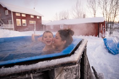 Ein Junge lächelt fröhlich, während er in einem heißen Whirlpool im Schnee sitzt, umgeben von einer winterlichen Landschaft.