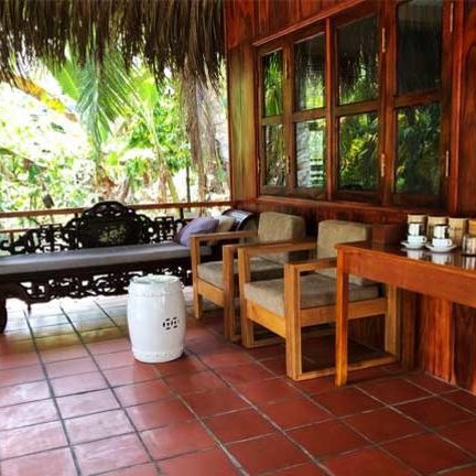 Eine gemütliche Terrasse mit Holzstühlen, einem Sofa und einem Tisch, umgeben von üppigem Grün und Palmen.