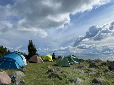 Eine Gruppe von Zelten steht auf einer Wiese, umgeben von Steinen, mit Blick auf einen ruhigen See und bewölkten Himmel.
