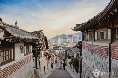Eine malerische Straße in Bukchon Hanok Village, gesäumt von traditionellen koreanischen Häusern und modernen Gebäuden im Hintergrund.