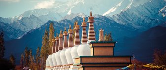Eine Reihe von Stupas mit goldenen Spitzen steht vor majestätischen, schneebedeckten Bergen im Hintergrund.