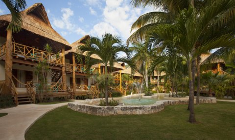 Ein tropischer Garten mit Palmen und einem kleinen Teich, umgeben von rustikalen Hütten mit Strohdächern.