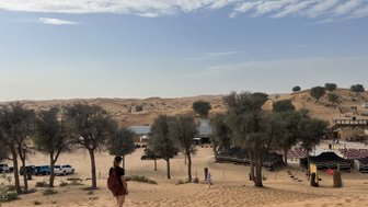 Eine Person mit einem roten Rucksack geht durch die Sanddünen, umgeben von Bäumen und einem Zeltlager im Hintergrund.