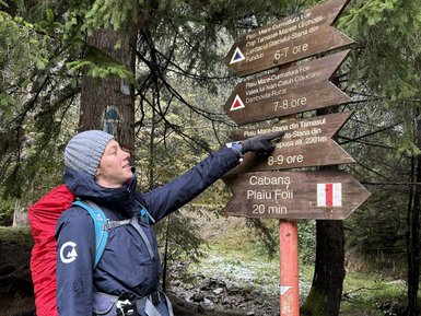 Eine Person in wetterfester Kleidung zeigt auf ein Wegweiser-Schild im Wald, umgeben von grünen Bäumen und einem kleinen Bach.