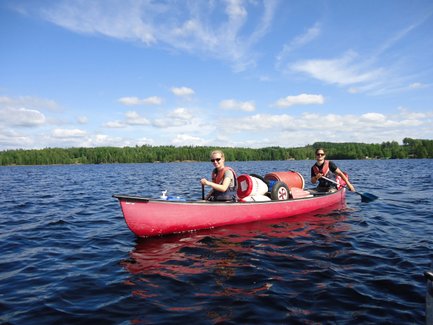 Zwei Personen paddeln in einem roten Kanu auf einem ruhigen See, umgeben von grünen Bäumen und blauem Himmel.