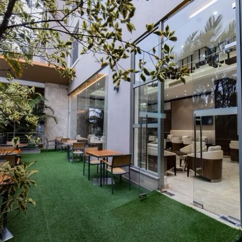 Ein einladender Außenbereich eines Restaurants mit Tischen und Stühlen, umgeben von üppigem Grün und modernen Fenstern.