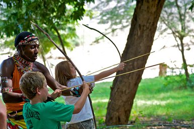 Ein Massai-Krieger zeigt zwei Kinder, wie man mit Pfeil und Bogen schießt, umgeben von üppiger Natur.