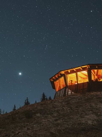 Eine beleuchtete Hütte steht auf einem Hügel, umgeben von dunklem Himmel und funkelnden Sternen.