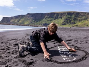 Ein Junge kniet am schwarzen Sandstrand und gestaltet mit Steinen eine kreative Form im Sand.