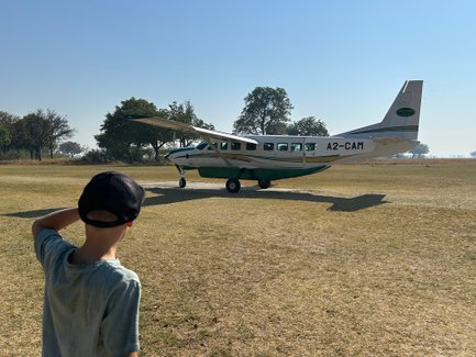 Ein Junge mit einer Kappe beobachtet ein kleines Flugzeug auf einem offenen Feld in Botswana unter klarem Himmel.