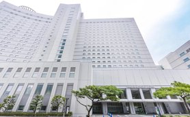 Ein modernes Hotelgebäude mit einer hellen Fassade und großen Fenstern, umgeben von gepflegten Bäumen und einem Parkplatz.