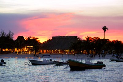 Ein malerischer Sonnenuntergang über einem ruhigen Gewässer, umgeben von Palmen und beleuchteten Restaurants am Strand.