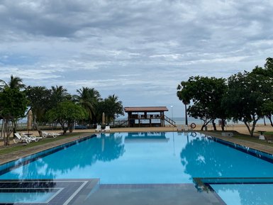 Ein weitläufiger Pool spiegelt den bewölkten Himmel wider, umgeben von Palmen und einer einladenden Liegewiese.