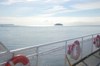 Ein Blick von einem Schiff auf das ruhige Wasser, mit einer kleinen Insel in der Ferne und einem klaren Himmel darüber.