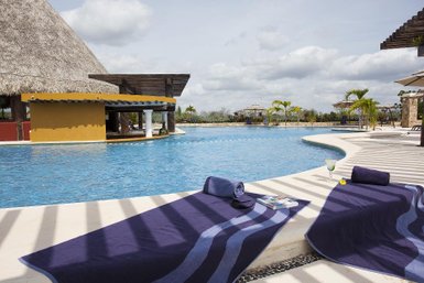 Ein einladender Poolbereich mit blauen Wasser, umgeben von Palmen und einem strohgedeckten Gebäude im Hintergrund.