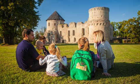 Eine Familie sitzt auf dem Gras vor einer alten Burg, während sie sich unterhält und die Sonne genießt.