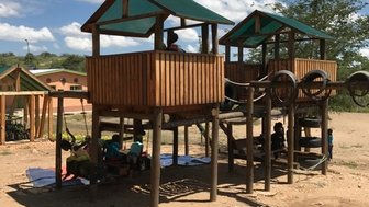 Ein Spielplatz mit zwei Holzhäusern auf Stelzen, umgeben von einem klaren blauen Himmel und sanften Wolken.