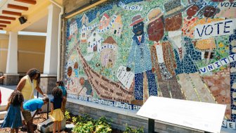 Eine Mutter hilft ihren Kindern, während sie vor einem bunten Mosaik mit historischen Themen steht, das Gemeinschaft und Einheit feiert.