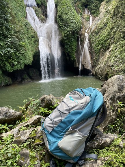 Ein blauer Rucksack steht auf einem Felsen, während ein Wasserfall im Hintergrund in einen klaren Teich fließt.