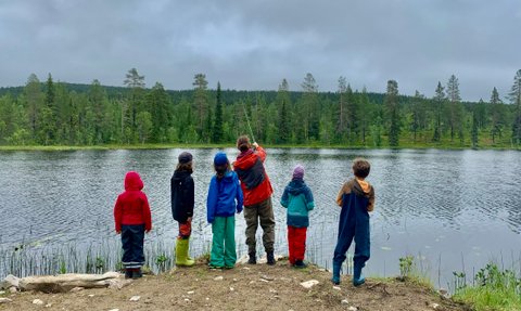 Sechs Kinder stehen am Ufer eines ruhigen Sees und beobachten einen Erwachsenen beim Angeln in der Natur.