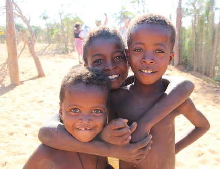Drei fröhliche Kinder umarmen sich in der Sonne, umgeben von einer sandigen Landschaft und grünen Pflanzen im Hintergrund.