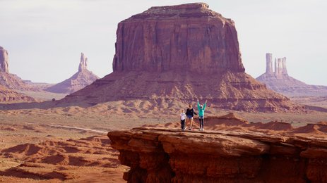 Drei Personen stehen auf einer Felsplattform in Monument Valley, umgeben von beeindruckenden roten Felsen und Hügeln.