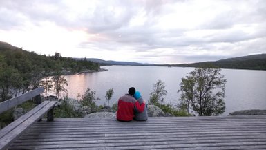Ein Paar sitzt auf einer Holzterrasse am See und genießt den Sonnenuntergang, umgeben von einer malerischen Landschaft.