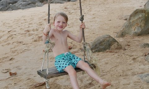 Ein fröhlicher Junge schwingt auf einer Holzschaukel am Strand, umgeben von einer malerischen Küstenlandschaft.