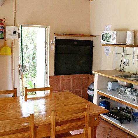 Eine helle Küche mit einem großen Holztisch, Kühlschrank und einer offenen Tür, die auf einen grünen Außenbereich führt.