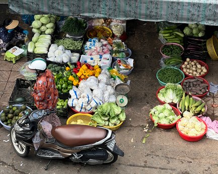 Ein belebter Marktstand in Saigon, voll mit frischem Gemüse und Obst, während eine Person in traditioneller Kleidung einkauft.