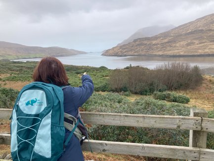 Eine Person mit einem Rucksack zeigt auf die ruhige Killary Fjord-Landschaft, umgeben von sanften Hügeln und bewaldeten Ufern.