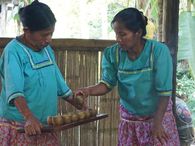 Zwei Frauen in traditioneller Kleidung bereiten gemeinsam Speisen in einer ländlichen Umgebung mit Holz und Töpfen vor.