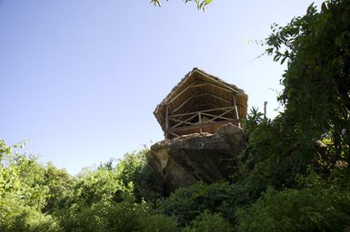 Eine Aussichtshütte aus Holz mit einem Strohdach, die auf einem großen Felsen inmitten üppiger Vegetation thront.