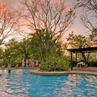 Ein einladender Pool umgeben von üppigem Grün und Bäumen, während die Sonne am Horizont untergeht.