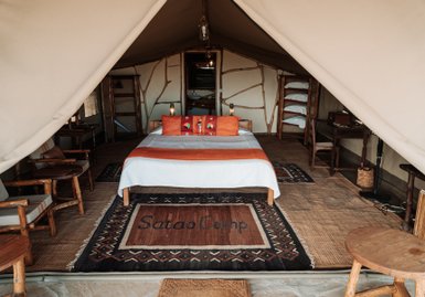 Ein gemütliches Zeltinnere mit einem großen Bett, bunten Kissen und einem Teppich mit der Aufschrift 'Satao Camp'.