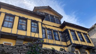 Ein historisches gelbes Gebäude mit kunstvollen Fenstern und einem steinernen Fundament, umgeben von üppigem Grün.